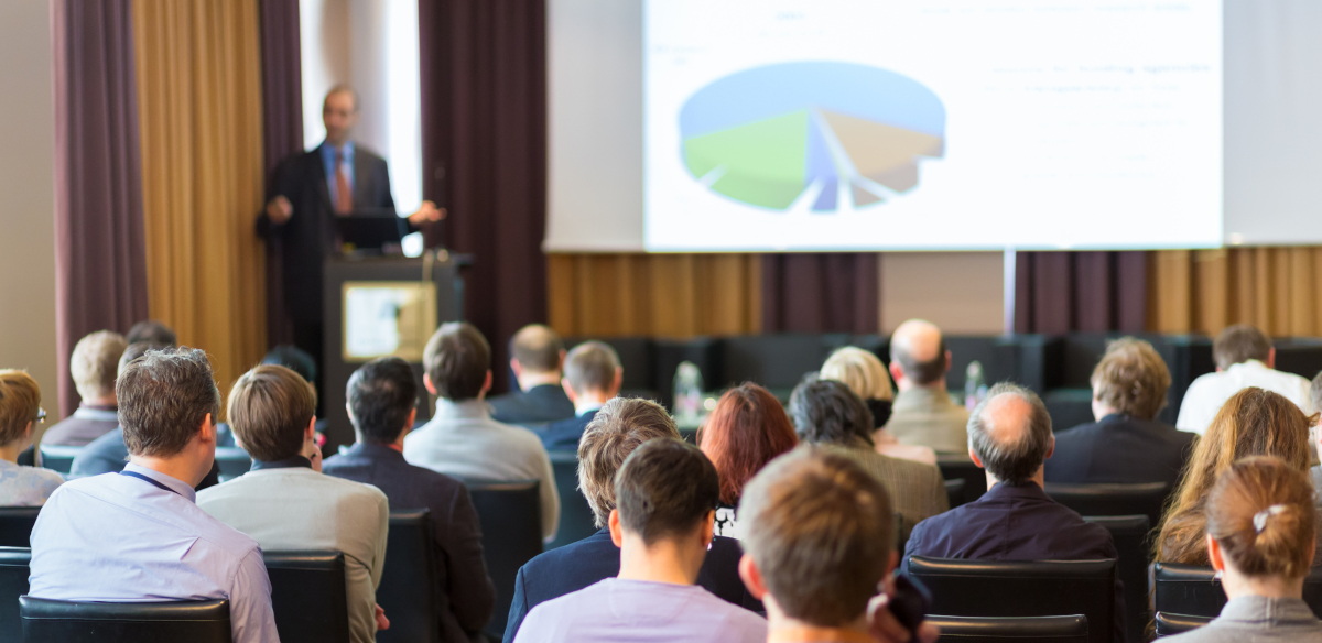 A moderately sized group of people attending a convention and listening to a speaker give a presentation.