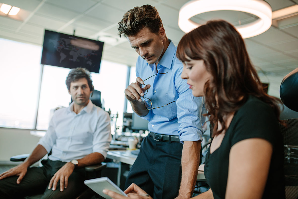 Mature businessman looking at notepad shown by female colleague with a man sitting in background. Business people working together in office.