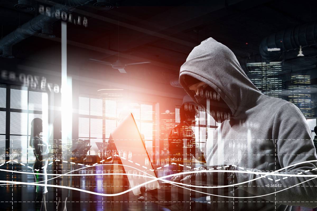 A hacker in a gray hooded sweatshirt and a face covering using a laptop to steal information