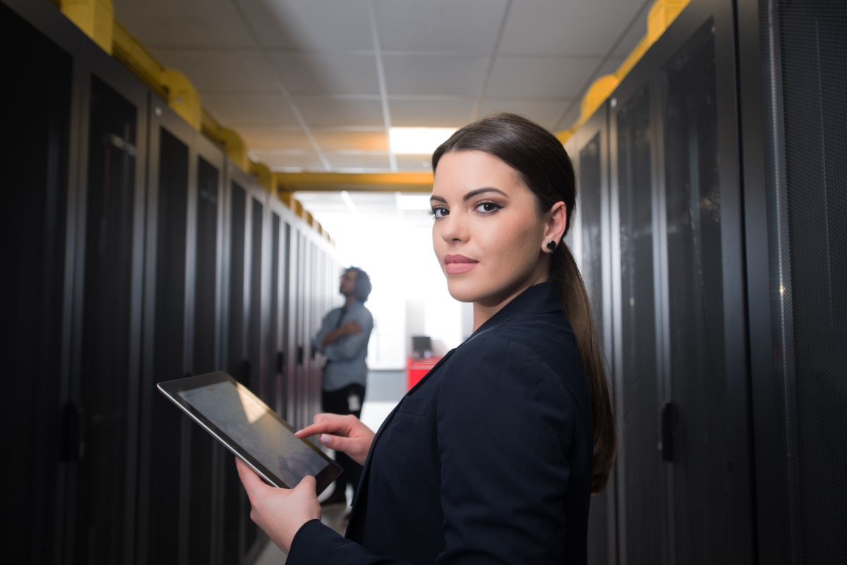A brunette woman standing in a server room using a laptop