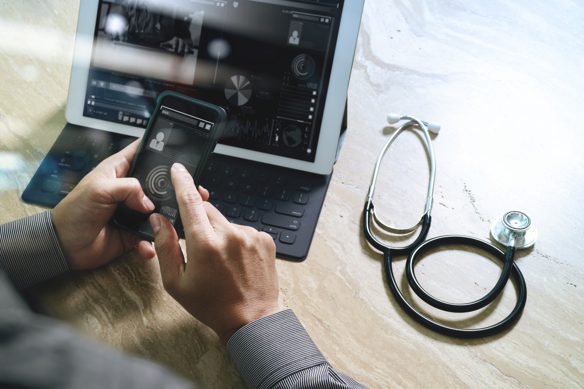 A man using a phone and laptop with a stethoscope on the desk next to his computer