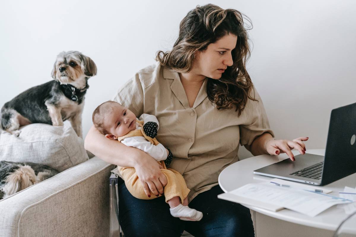 A woman working from home while holding a baby and two dogs are on the couch behind her