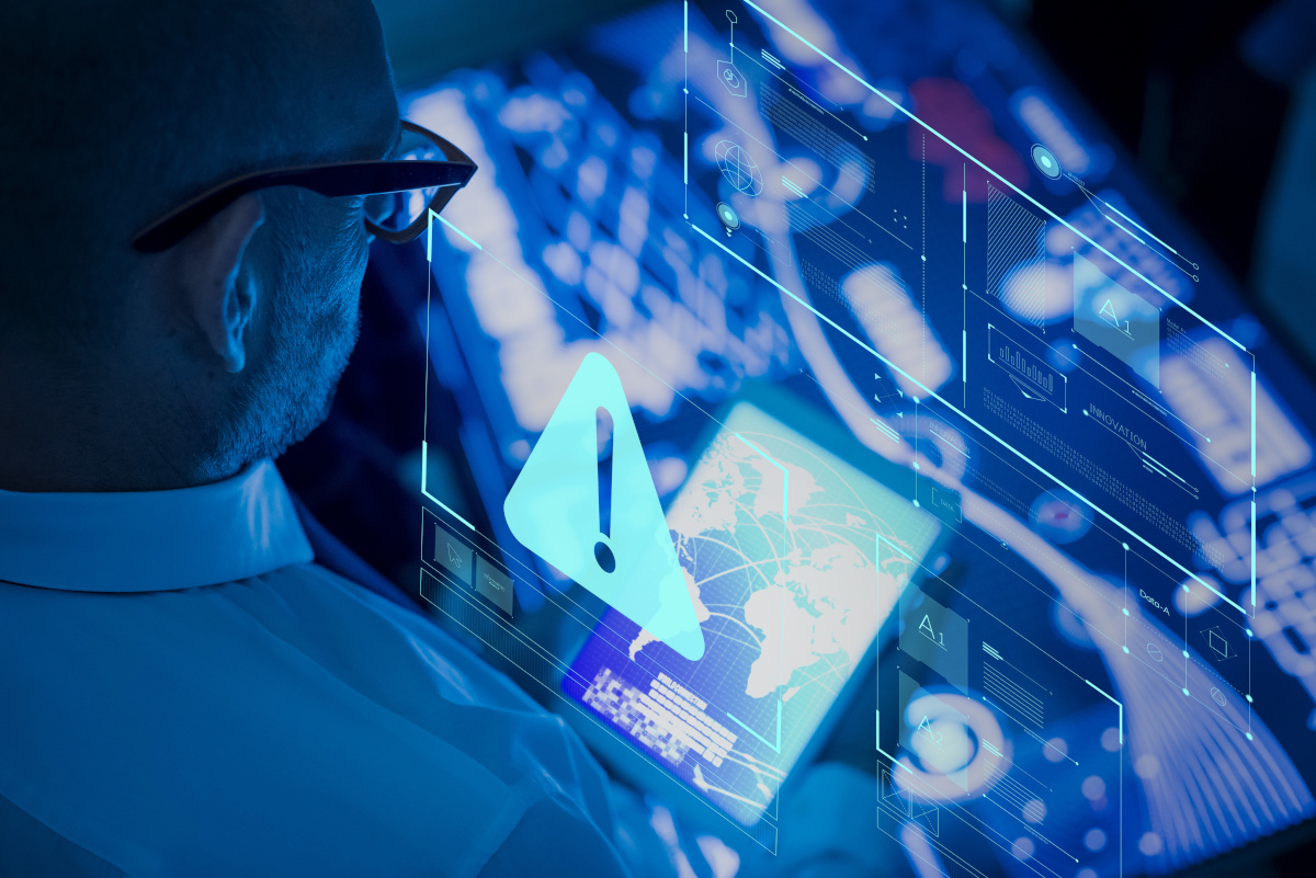 A man looking at a tablet as it displays a world map, while also presenting a holographic display with a warning symbol in front of him