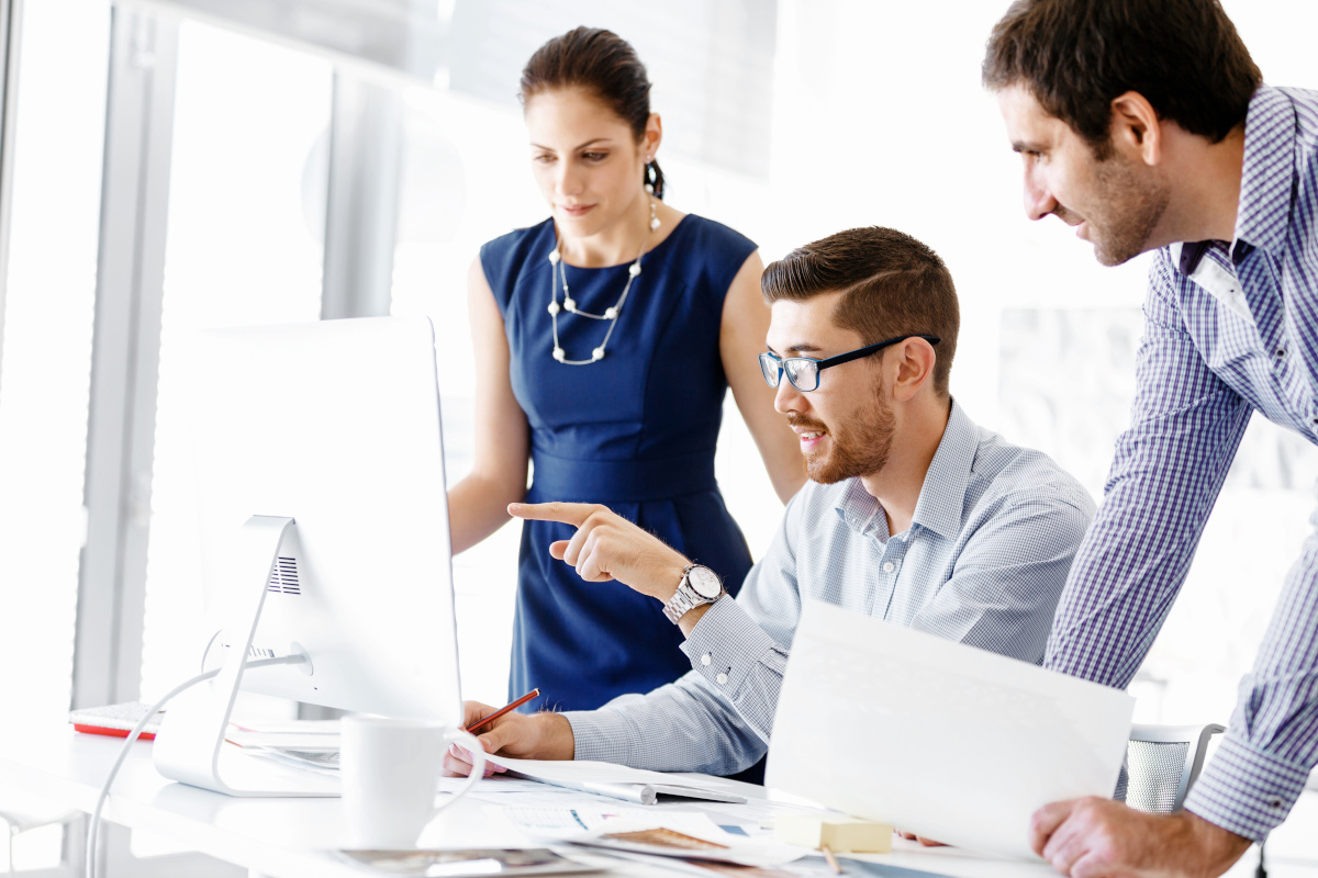 Three people looking at a computer monitor while the one sitting at the desk points towards the screen