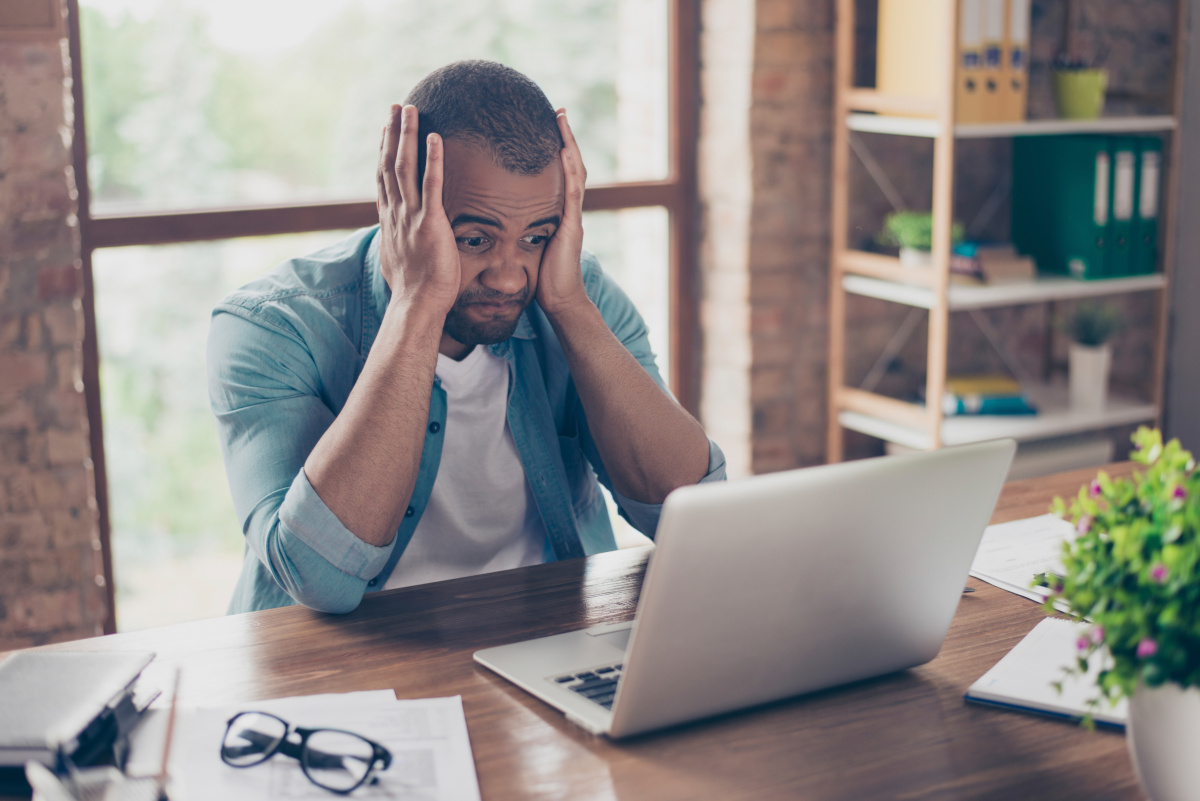 A man sandwiching his head between his hand with a clear look of frustration while sitting at his home office desk staring at his laptop