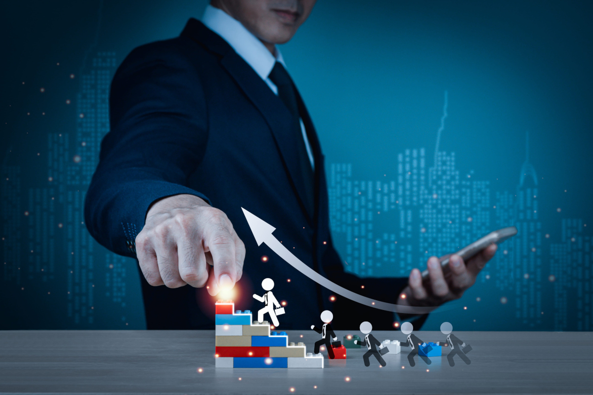A businessman placing a glowing light piece on top of a toy block staircase as small illustrated business figures climb upward toward a rising arrow.