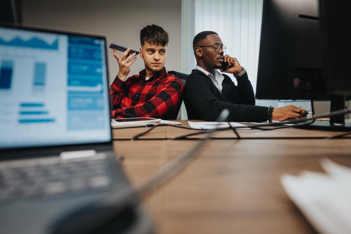 Two employees at office desks taking calls beside laptops and a monitor.
